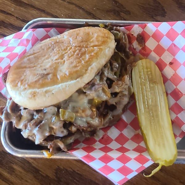 Longhorn Outpost & Barbeque Company's cheesesteak sandwich with a pickle spear on a checkered paper-lined tray.