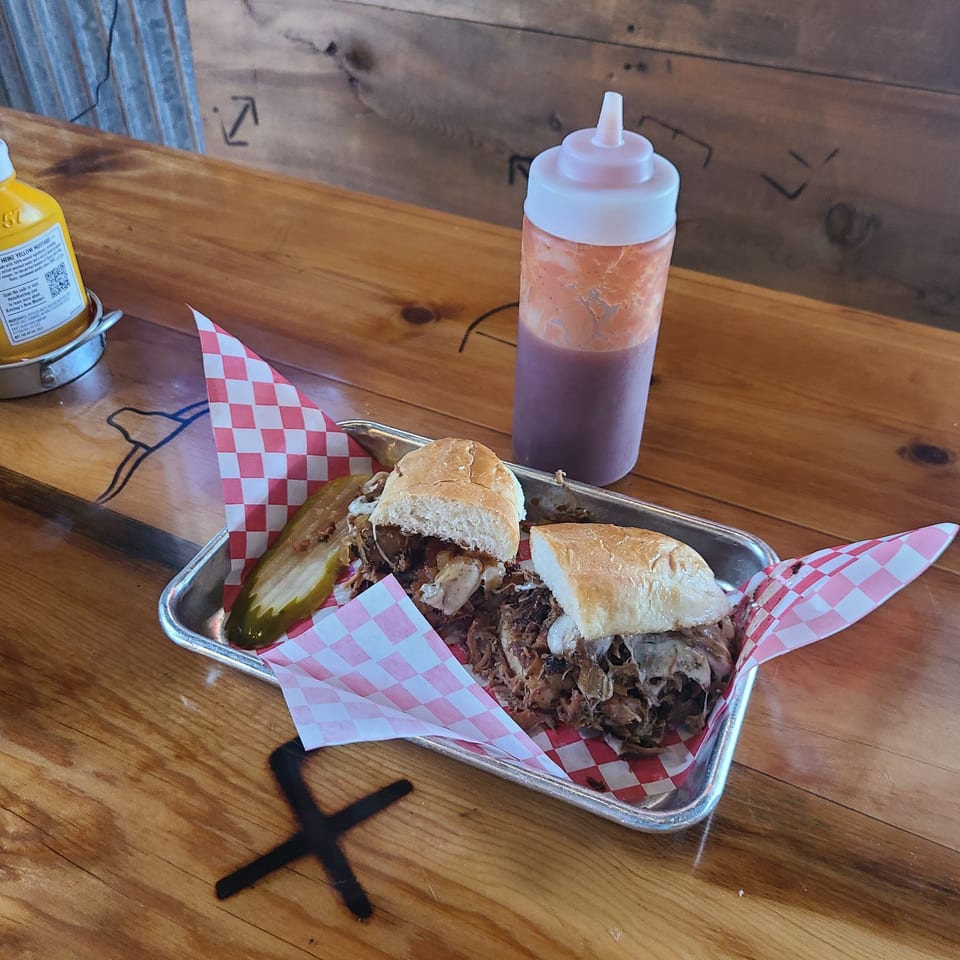A Longhorn Outpost & Barbeque Company pulled pork sandwich with a pickle spear and sauce bottles.